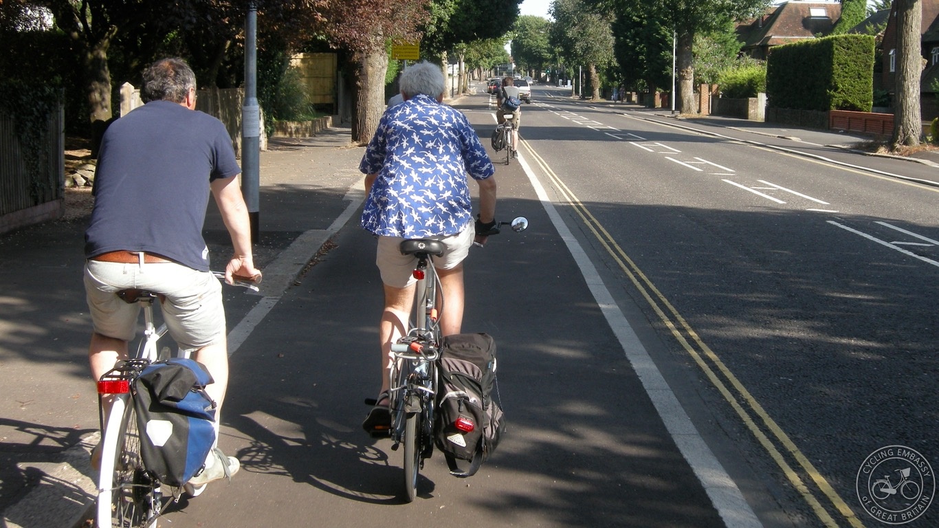 Photo showing a two-way road with a footway to its left, and a separate wide cycle track between the road and the footway, with kerbstones at the edges of both the footway and the cycle track (Source: Cycling Embassy of Great Britain https://www.cycling-embassy.org.uk/dictionary/hybrid-cycle-track)
