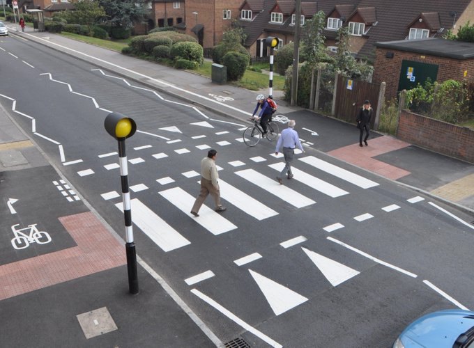 An image of a two-way road with a black and white striped zebra crossing (with Belisha beacons (a striped black and white pole with a yellow light at the top)) across it for pedestrians, with a separate, dotted-edge, crossing across it for cyclists; the whole dual crossing is raised, with triangular markings indicating the hump to vehicle drivers (Source: Essex Active Travel Design Portal https://www.cycling-embassy.org.uk/dictionary/hybrid-cycle-track)